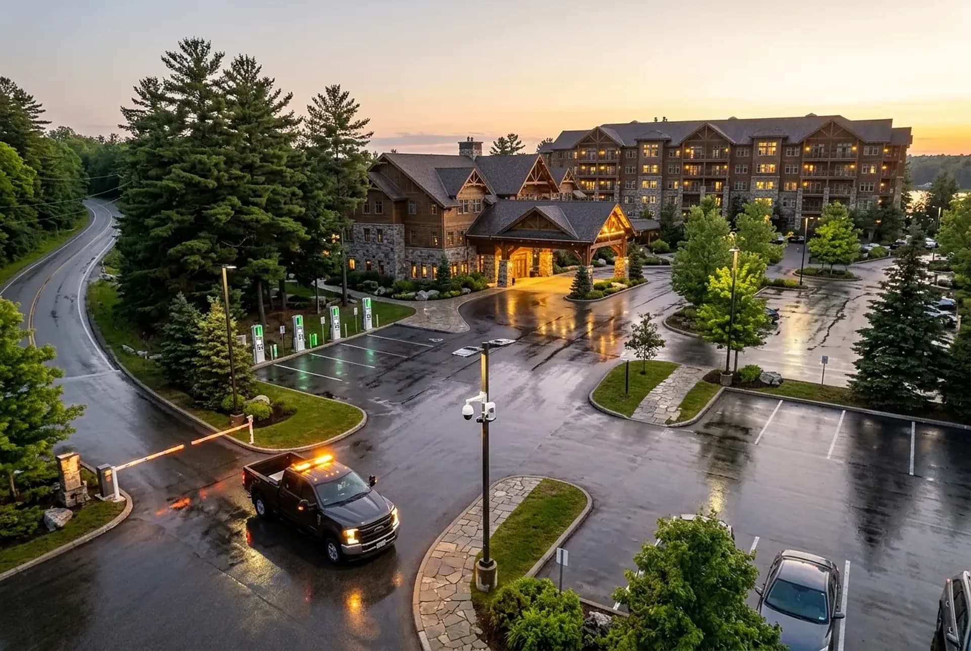 A hotel parking lot at dusk after a summer shower, a grounds pickup at the gate, a camera on a lamp post and the lit inn behind